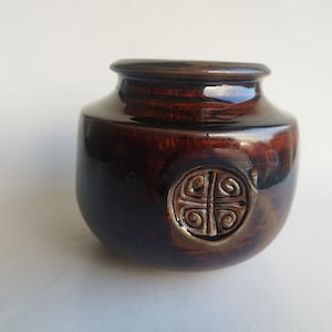 May include: A small, round, brown ceramic vase with a glossy finish. The vase features a decorative, embossed design on the front. The vase has a wide body and a narrow opening, suitable for small floral arrangements or decorative use.