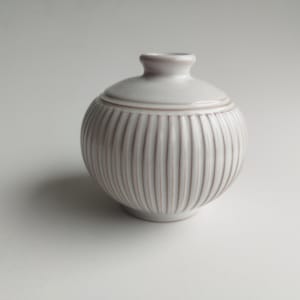 Puede incluir: Un pequeño tarro de cerámica blanco con tapa. El tarro tiene un cuerpo redondo con detalles acanalados verticales y un cuello pequeño y cilíndrico. La tapa también es blanca y tiene una forma similar al tarro. El tarro es probablemente para almacenamiento o decoración.
