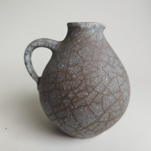 Könnte beinhalten: Ein kleiner, grauer Keramik Krug mit einer craquelé Glasur und einem Henkel. Der Krug hat einen runden Körper und eine leicht ausgestellte Lippe.