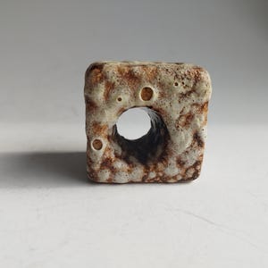 May include: A square, ceramic vase with a large circular opening. The vase has a textured, speckled surface with a mix of beige and brown tones. The design features several small, round indentations.