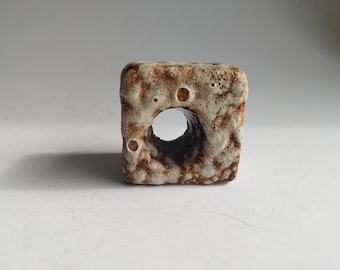 May include: A square, ceramic vase with a large circular opening. The vase has a textured, speckled surface with a mix of beige and brown tones. The design features several small, round indentations.