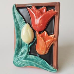 May include: A ceramic tile with a raised design of three tulips. The tulips are orange, red, and white. The tile is framed in a terracotta border.