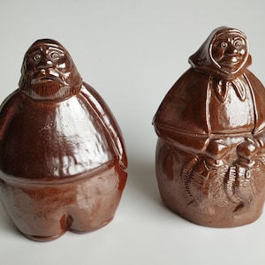 May include: Two ceramic figurines, one depicting a man and the other a woman. The man is wearing a brown hat and coat, while the woman is wearing a brown hooded cloak and holding two fish. Both figurines are brown and have a glazed finish.