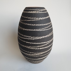 Puede incluir: Un jarrón de cerámica negro con rayas horizontales blancas. El jarrón tiene forma ovalada y una superficie lisa y texturizada.
