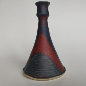 May include: A ceramic candlestick holder with a flared base and a narrow neck. The surface features a matte dark blue and reddish-brown color scheme with a textured, ribbed pattern around the lower section and a smooth finish at the top.