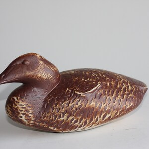 May include: A brown ceramic duck figurine with a detailed textured surface. The duck has a long, thin beak and a rounded body.