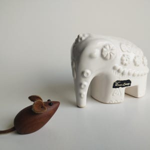 Puede incluir: Una figurita de elefante de cerámica blanca con intrincados diseños florales y una etiqueta negra que dice "Rosa Ljung" está al lado de una figurita de ratón de madera con orejas de piel marrón.