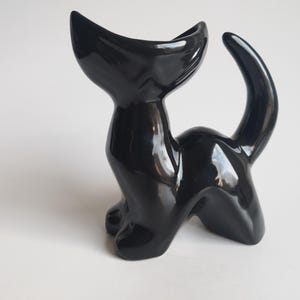 May include: A shiny black ceramic cat ornament with a curved tail and a wide opening at the top. The cat is in a seated position, with its front paws visible. The ornament has a smooth, reflective surface.