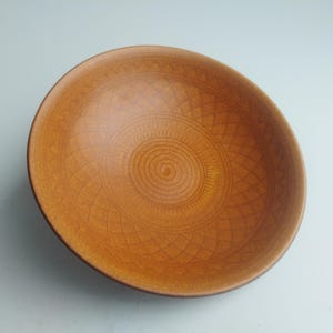 May include: A round, brown ceramic bowl with an intricate, geometric pattern etched into the interior. The bowl's design features concentric circles and diamond shapes, creating a textured surface. The bowl is a warm, earthy tone.