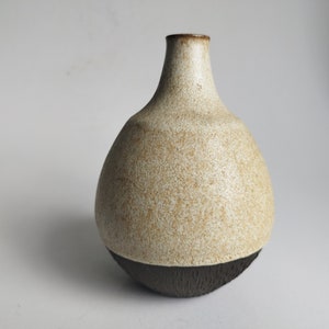 Puede incluir: Un pequeño jarrón de cerámica blanca con un esmalte moteado y una base marrón oscuro. El jarrón tiene un cuerpo redondeado y un cuello estrecho.