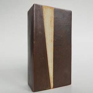 Pode incluir: Um vaso retangular de cerâmica com acabamento texturizado em castanho escuro. A superfície frontal apresenta uma incrustação triangular vertical em branco sujo que se estreita em direção à base, criando um contraste com o corpo em cerâmica.