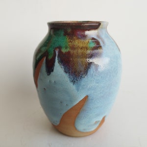 May include: A small, hand-thrown ceramic vase with a blue glaze and brown, green, and purple accents. The vase has an irregular shape and a slightly flared opening.
