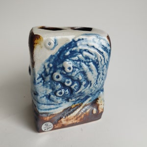 May include: A white ceramic toothbrush holder with blue and brown glaze. The holder has a textured surface and a unique design. It is signed "S. Jackson" on the bottom.