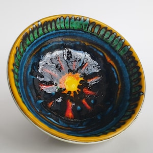 May include: A ceramic bowl with a vibrant, abstract design. The bowl features a yellow rim, a green and blue border, and a dark interior with orange and yellow accents. The bowl is likely used for serving food or as a decorative piece.