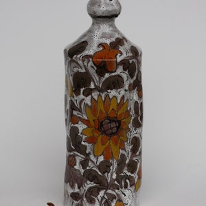 May include: A white ceramic vase with a floral design. The vase has a wide mouth and a narrow neck. The flowers are orange, yellow, and brown. The vase is decorated with a white glaze.