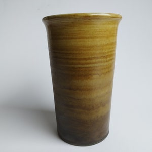 May include: A tall, cylindrical ceramic vase with a brown and yellow glaze. The glaze has a streaky, layered effect, creating a gradient from light yellow at the top to dark brown at the bottom.