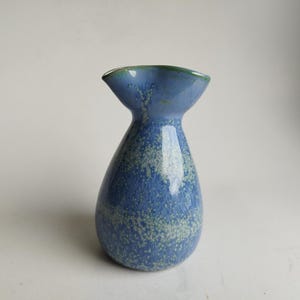 May include: A blue ceramic vase with a distinctive form. The vase features a broad, flared top and a rounded body. The blue glaze is speckled with lighter blue and green, giving a textured look. The rim is a darker green.