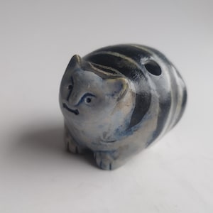 May include: A ceramic cat figurine with a blue and white color scheme. The cat has a striped pattern on its back and a hole on top. The figurine has a grumpy expression and is sitting on its four paws.