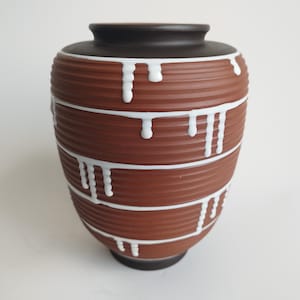 Puede incluir: Un jarrón de cerámica marrón con borde y base marrón oscuro. El jarrón tiene estrías horizontales y detalles de pintura blanca goteando. Es un objeto decorativo, adecuado para exhibir flores o como pieza de arte independiente.