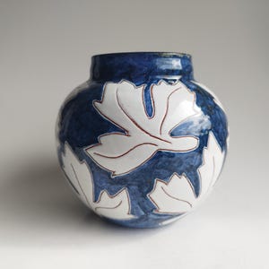 Puede incluir: Un jarrón de cerámica redondo con fondo azul oscuro y diseños de hojas blancas delineadas en rojo. El jarrón tiene un cuerpo ancho y un cuello estrecho, adecuado para la decoración del hogar.
