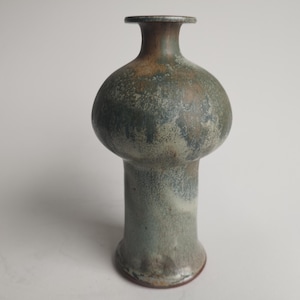 Puede incluir: Un jarrón de cerámica con un cuerpo bulboso y un cuello alto y delgado. El jarrón está esmaltado en tonos verdes y marrones, con un acabado moteado.