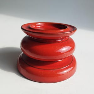 Könnte beinhalten: Ein glänzender, roter Keramik-Kerzenhalter mit einem mehrschichtigen Design. Der Halter hat eine kreisförmige Basis, einen abgerundeten Mittelteil und eine Oberseite mit einem kleinen, vertieften Bereich. Die Gesamtform erinnert an eine gestapelte Form.