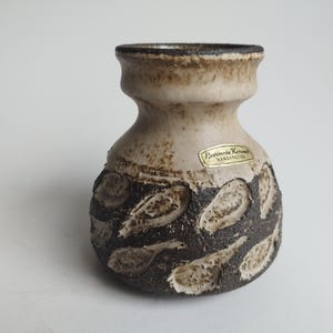 May include: A small, white ceramic vase with a brown and white speckled glaze. The vase has a rounded body and a narrow neck. It is decorated with a pattern of brown leaves. The vase has a label that reads "Bernermode Keramik Handarbeit".