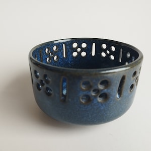 May include: A small, round, ceramic bowl with a dark blue glaze. The bowl features a series of decorative cut-out holes around the rim. The holes are arranged in a pattern of circles and vertical lines. The bowl is likely used for decorative purposes.