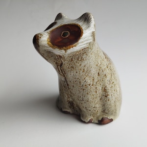 Könnte beinhalten: Keramik-Waschbärfigur mit strukturiertem, gesprenkeltem Körper in Beige- und Brauntönen. Der Waschbär hat eine markante braune Maske um die Augen und eine kleine, hochgezogene Nase. Dekorativer Artikel.