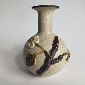 May include: A small, cream-colored ceramic vase with a brown speckled glaze. The vase has a round body and a narrow neck. It is decorated with brown and white raised floral designs.
