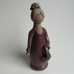 Puede incluir: Una figurita de cerámica de una mujer con un moño en el pelo, vestida con un vestido morado y sosteniendo una cesta.