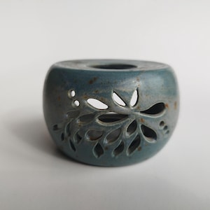 May include: A teal ceramic candle holder with a floral cutout design. The round holder has a small opening at the top and a series of leaf-shaped cutouts on the side. The ceramic has a speckled texture.