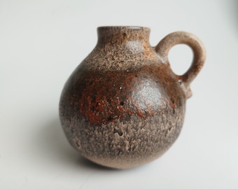 May include: A small, brown and gray ceramic vase with a textured surface and a single handle. The vase has a rounded body and a slightly flared opening.