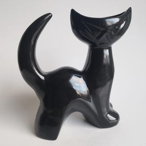 May include: A black ceramic cat figurine with a curled tail and a stylised face. The cat is standing on all four legs and has a glossy finish.