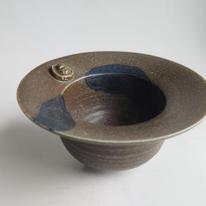 May include: A handmade ceramic bowl with a wide, flat rim and a deep, rounded interior. The bowl is primarily brown with blue accents and a small, raised decorative element on the rim. The bowl's diameter is approximately 8 inches.
