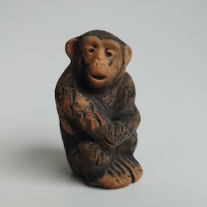 May include: A small, brown ceramic monkey figurine with a detailed face and a seated pose. The monkey's arms are crossed and its legs are bent.