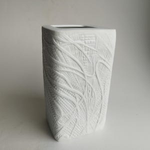 Op de afbeelding: Een witte keramische vaas met een vierkante bovenkant en een getextureerde, patroonrijke buitenkant. Het patroon heeft een netwerk van lijnen en vormen, waardoor een gedetailleerd, artistiek ontwerp ontstaat. De vaas heeft een moderne, geometrische vorm.