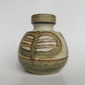 Puede incluir: Un pequeño jarrón de cerámica redondo con cuello estrecho y cuerpo ancho y bulboso. La superficie presenta un esmalte moteado en tonos beige tierra y verde oliva con bandas horizontales de color marrón oscuro y un patrón simétrico de hojas.