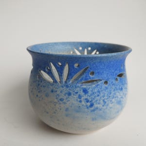 May include: A small, blue and white ceramic candle holder with a decorative cut-out design. The holder has a speckled glaze and a rounded shape.
