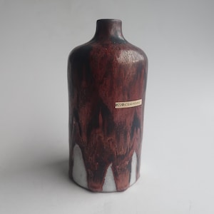 Könnte beinhalten: Eine Keramikvase mit dunkelroter und schwarzer Glasur, mit einem Tropfmuster, das an der Basis zu Weiß verblasst. Die Vase hat einen schmalen Hals und ein Etikett mit der Aufschrift "DZO KERAMIK".