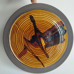 May include: A large, round ceramic plate with a dark brown rim and a concentric circle design in shades of brown and black. The plate has a unique abstract design in black and brown.