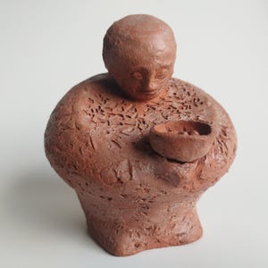 May include: A terracotta sculpture of a person holding a bowl. The sculpture is detailed with textured lines and patterns.