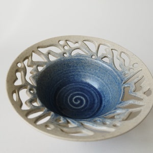 Pode incluir: Tigela de cerâmica com exterior bege claro e interior azul profundo. A borda apresenta um design decorativo recortado. O interior da tigela tem um padrão em espiral. Uma peça decorativa para a casa.