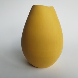 Puede incluir: Un jarrón amarillo con un acabado liso y mate. El jarrón tiene una forma redondeada de lágrima con una abertura ligeramente ensanchada en la parte superior. El jarrón mide aproximadamente 30 cm de alto y 18 cm de ancho.