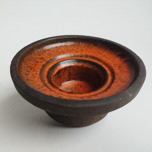 May include: A ceramic candle holder with a dark brown base and rim, and a vibrant orange interior. The interior has a textured, crackled appearance. The candle holder has a circular shape with a recessed center.