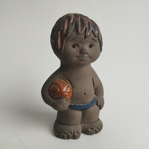 May include: A brown ceramic figurine of a child holding an orange ball. The child is wearing blue swim trunks and has brown hair.