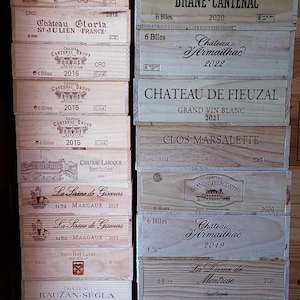 May include: A stack of wooden wine boxes with labels featuring the names of different French wineries, including Chateau Larogue, Chateau Siran, Clos Marsalette, Chateau Gloria, Chateau Cantenac Brown, Chateau Armailhac, Chateau de Fieuzal, Grand Puy Lacoste, La Sirene de Giscours, Rauzan Segla, and Chateau d'Issan. The boxes are stacked in two columns, with the labels facing forward.