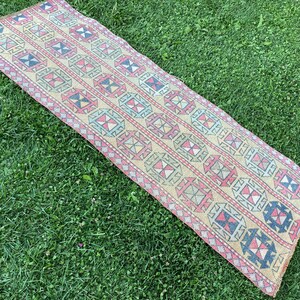 May include: A long, narrow rug with a geometric pattern in shades of pink, blue, yellow, and black. The rug is woven with a traditional design and has a fringed edge.