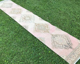 May include: A long, pink and cream runner rug with a diamond pattern and a fringe border. The rug is laid out on a green grassy lawn.