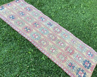 May include: A long, narrow rug with a geometric pattern in shades of pink, blue, yellow, and black. The rug is woven with a traditional design and has a fringed edge.